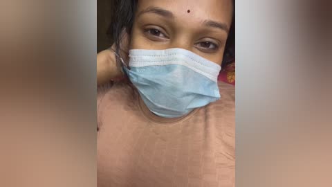 A close-up video of a South Asian woman with medium-brown skin, wearing a light blue medical mask, brown eyes, and short black hair. She has a mole on her forehead and is wearing a plain, light-colored shirt.