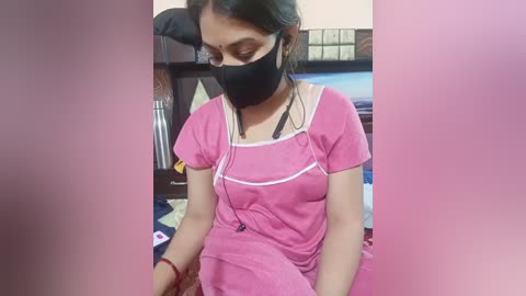 A video of a woman with medium skin tone, wearing a pink t-shirt, black mask, and headphones, sitting on a bed with pink sheets.