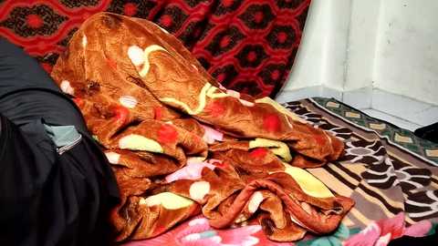 Video of a person lying on a bed, covered with a colorful, patterned blanket. The background features traditional red and black patterned textiles. The setting suggests a warm, cozy environment.