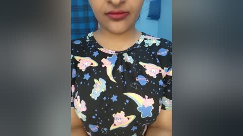 Video of a woman with light brown skin, wearing a black top with colorful, playful cartoon space-themed prints, including a pink unicorn, a blue rocket, and a yellow crescent moon.