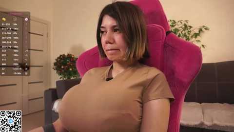 A photo of a woman with a large, exaggeratedly pregnant belly in a pink gaming chair, wearing a beige shirt. Background includes a floral arrangement, beige walls, and a gray couch.