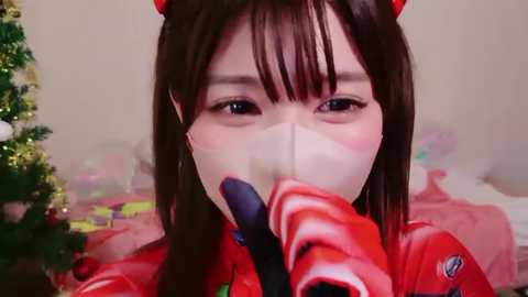 Video of a young woman wearing a red and white striped face mask, black gloves, and a red and white striped jacket, with red cat ears, in a festive, decorated room.
