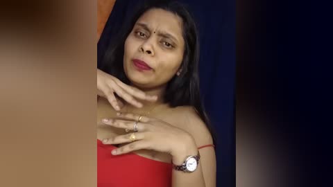 Video of a South Asian woman with medium brown skin, straight black hair, wearing a red off-shoulder top, with a red bindi on her forehead. She has a neutral expression and touches her neck with her right hand.