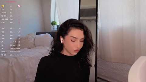 Media: Video of a young woman with curly black hair, wearing a black top, sitting on a beige couch in a minimalist, well-lit room.