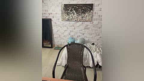 Video of a minimalist bedroom with a white brick wall, a black-framed mirror, and a wicker chair facing a bed with white linens and two blue pillows.