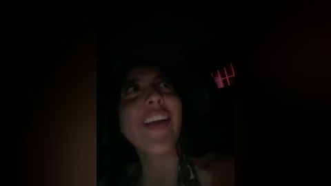 A dimly lit video of a joyful woman with medium-dark skin and curly hair, wearing a green top, smiling widely. A red window light illuminates her face.