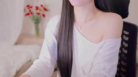 A video of a young woman with long, straight black hair, wearing a white off-shoulder blouse, seated in a modern bedroom with white bedding and a vase of red flowers.