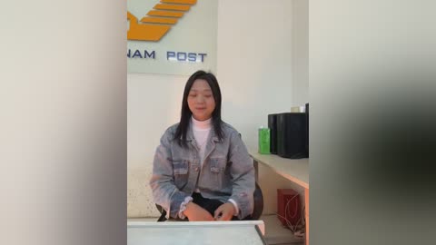 A video of an Asian woman with long black hair and fair skin, sitting at a desk in a modern office. She wears a white turtleneck under a denim jacket. The background includes a yellow staircase, a green plant, and a black monitor.