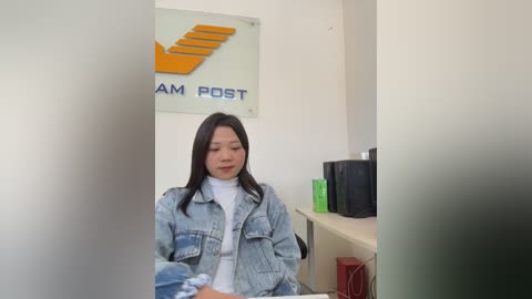 A video of an Asian woman with long black hair, wearing a denim jacket over a white turtleneck, seated in a modern office with white walls, a large \"AM POST\" sign, and office equipment.