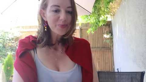Media: Video of a fair-skinned, light-haired woman with a red shawl draped over her shoulders, wearing a white tank top, smiling outdoors near a wooden fence and greenery.