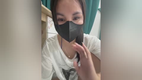 Media: Video of an Asian woman with straight black hair, wearing a black face mask and white t-shirt, seated in a bathroom with teal curtains and a wooden shelf.