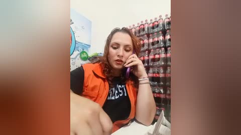 A video of a fair-skinned woman with long brown hair, wearing an orange vest, talking on a phone while seated in a store with shelves of bottled drinks in the background.