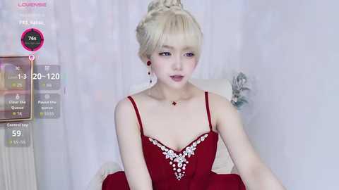 Video of a young woman with platinum blonde hair in an updo, wearing a red lace dress with white floral embroidery. She sits in a room with white curtains.