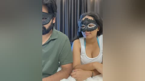 Video of a South Asian couple wearing black Venetian masks, the man in a green polo and the woman in a white tank top, sitting close on a fluffy white blanket.