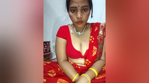 Video of a young South Asian woman with medium skin tone, wearing a red sari with yellow patterns, adorned with gold jewelry, seated indoors against a white wall.