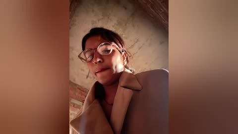 A video captures a young woman with short brown hair, wearing glasses and a beige coat, sitting in an old, brick-walled room with a textured ceiling. The lighting is soft and warm.