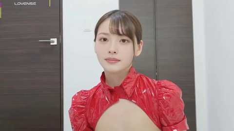 Video of a young East Asian woman with straight, dark brown hair, wearing a shiny red, puffed-sleeve blouse with a deep neckline, standing in front of dark wooden doors.