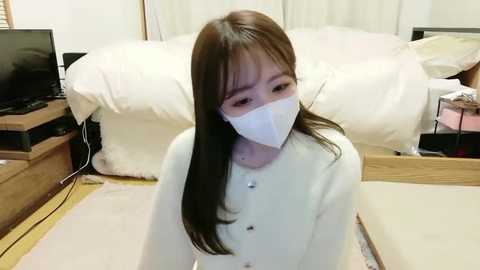 Video of an Asian woman with long brown hair, wearing a white face mask, white cardigan, and beige pants, kneeling on a light carpet in a traditional Japanese room with tatami mats, wooden furniture, and a white futon bed.
