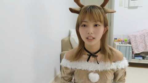 Media: Video of an Asian woman with light skin, wearing a brown off-shoulder velvet dress with white fur trim, and brown antler headband, in a simple, beige room.