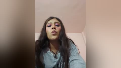 Video of a young woman with long dark hair, wearing a light blue shirt, standing against a beige wall. She has a neutral expression, slightly puckered lips, and light makeup.