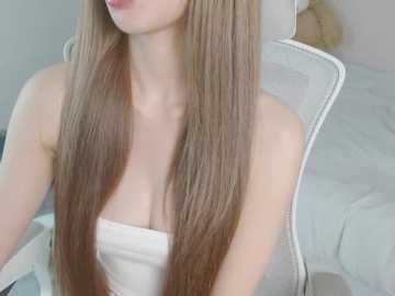 Video of a fair-skinned woman with long, straight, light brown hair, wearing a strapless white top, sitting on a white chair in a softly lit, minimalist room.