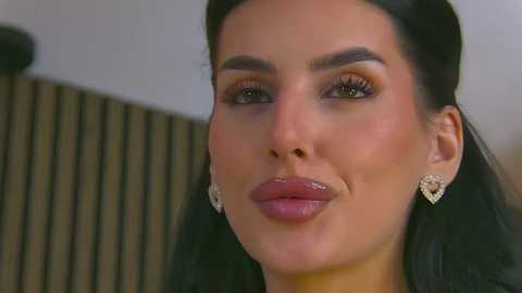 Video of a woman with dark hair, fair skin, and full lips, wearing large heart-shaped earrings, and a dark outfit, against a blurred striped background.