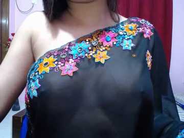 Video of a woman with medium skin tone wearing a black, sheer, one-shoulder top adorned with colorful floral embroidery, featuring blue, yellow, and pink flowers. Background includes a red curtain and pink wall.