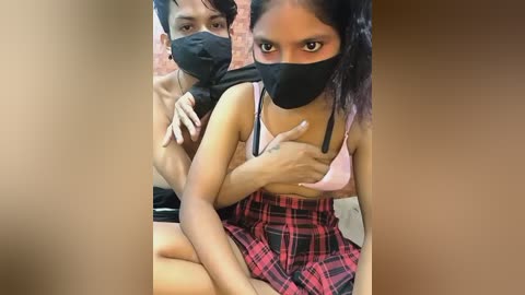 Video of two young, light-skinned South Asian women wearing black face masks, one topless, the other in a plaid skirt, sitting on a bed.