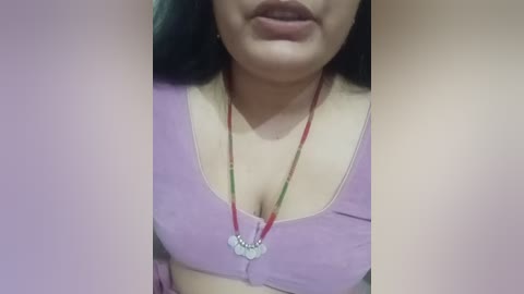 Video of a woman with medium skin tone, wearing a purple bra with a plunging neckline and a beaded necklace. Her lips are slightly parted, and her face is partially visible.