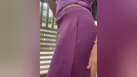 Media: Video of a person wearing a tight, purple, high-waisted skirt, captured from a low angle, showcasing their curved buttocks. The background reveals a window with white blinds and a glimpse of greenery outside.