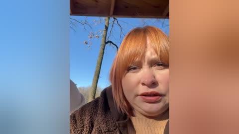 Video of a fair-skinned woman with short, orange-brown hair, wearing a brown coat, standing outdoors with a clear blue sky and leafless trees in the background.