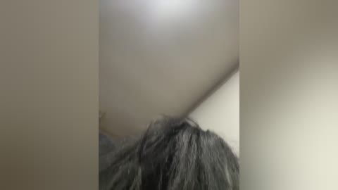 A video showing the upper part of a person's head with black, wavy hair. The image is blurry, focusing on the hair, with a white ceiling in the background. The lighting is soft, creating a muted, diffused effect.