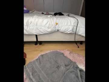 A video shows a messy bedroom with an unmade bed, a cluttered floor, and a gray and pink shaggy rug.