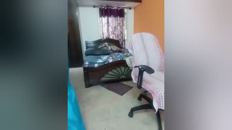 A video of a sparsely furnished bedroom with a wooden bed, blue floral sheets, and a white, patterned chair. The room has beige walls, a brown curtain, and a pink and white rug.