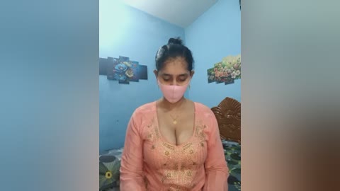 Video of a woman in a pink kurta, face mask, and bun hairstyle, standing in a blue-walled room with floral artwork, and a bed with green and brown patterned sheets.