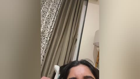 A video shows a partially visible person with black hair, holding a white toothbrush, standing in a beige bathroom with patterned curtains and a lamp.