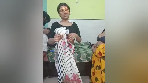 Video of a woman with a white handkerchief stained with blood, wearing a black top, standing in a room with a camouflage-patterned bed and a yellow wall.