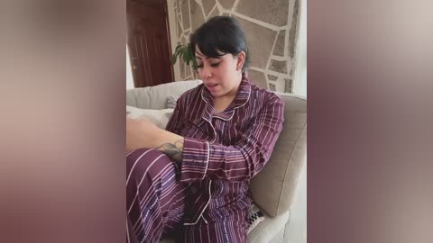 Video of a young woman with short black hair, wearing maroon striped pajamas, sitting on a beige couch, indoors, with a stone wall and wooden door in the background.