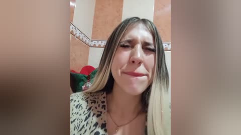 Video of a young woman with long, straight blonde hair, wearing a leopard print top, standing in a bathroom with beige and brown tiled walls and a red bathrobe on the left. She has a sad expression.