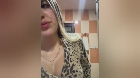 Video of a young woman with long, straight blonde hair, wearing a leopard print dress, standing in a bathroom with a pink and beige tiled wall.