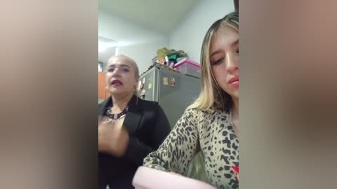 Video of a young woman with fair skin, dark hair, and a leopard print top, looking downcast. Behind her, a man with light skin, bald head, and a black shirt stands, seemingly unaware of her distress.