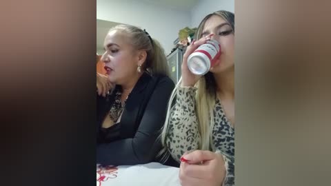 Video of two young women drinking from red and white soda cans in a dimly lit, indoor setting. One woman has light skin, blonde hair, and wears a black jacket. The other has tanned skin, long blonde hair, and a leopard print cardigan.
