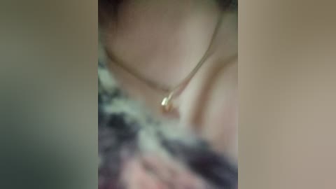 A blurry video of a person's neck and chest, showing a delicate gold necklace with a small pendant. The individual's skin appears fair. The background is out of focus.
