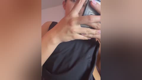Video of a woman with light skin, wearing a black sleeveless top, taking a mirror selfie with her left hand. Background is blurred, suggesting an indoor setting.