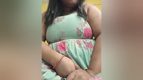 Video of a South Asian woman with medium skin tone and dark hair, wearing a turquoise floral dress, seated indoors with a yellow wall background.