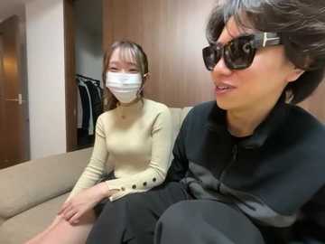 Media: Video of an Asian woman with short hair, wearing sunglasses and a mask, seated on a beige couch. Behind her, a woman with long hair in a beige mask and sweater is seated.