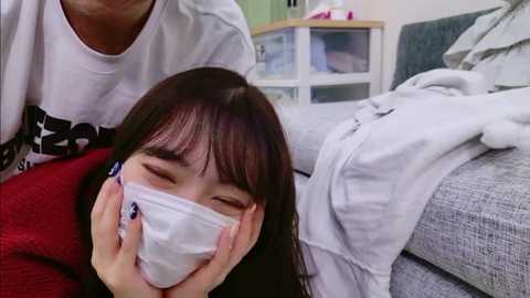 A video shows a young Asian woman with long black hair, wearing a white face mask, lying on a bed in a messy room.