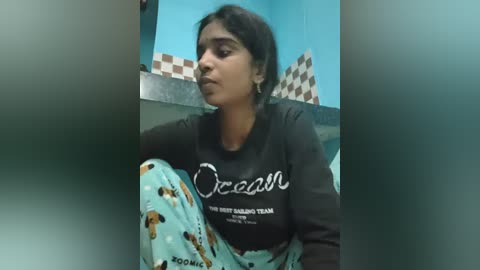 Video of a young woman with medium skin tone, dark hair, and brown eyes, wearing a black sweatshirt with \"Ocean\" and \"Oceana\" in white letters, and a patterned blanket. She sits on a teal-colored toilet seat in a restroom with blue and checkered wall tiles.