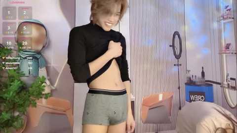 Video of a smiling, slender, light-skinned man with short, tousled hair, lifting a black shirt to reveal his toned abdomen. He wears gray athletic shorts. Background includes a green plant, a wall with a VR headset, and a desk with a computer monitor.