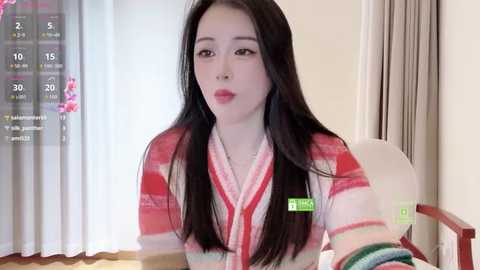 Video of an East Asian woman with long black hair, wearing a red and white striped kimono, seated at a table in a room with light curtains.